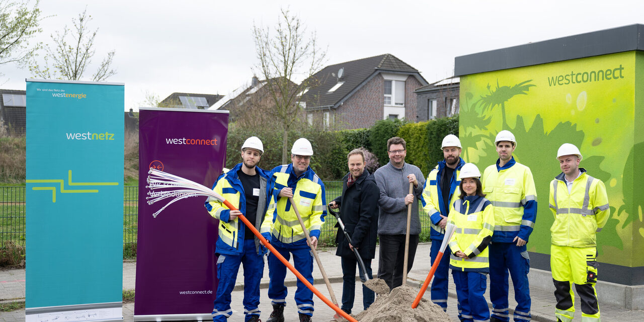 Mehrere Personen stehen um einen Sandhaufen und Glasfaserkabel herum. Im Hintergrund mehrere Banner von Westconnect
