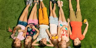 7 Kinder liegen auf einer Wiese und sind von oben aufgenommen worden. Sie tragen alle bunte Sommerkleidung und halten Obst oder Getränke in der Hand.