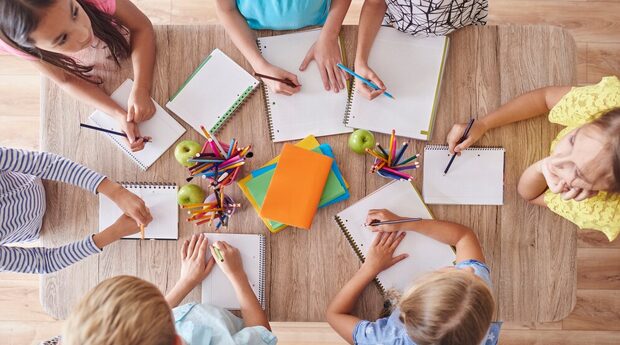 7 Kinder an einem Tisch, von oben fotografiert. Sie haben alle einen Block vor sich und einen Stift in der Hand. In der Mitte liegeen bunte Blätter, Äpfel und Stiftboxen.