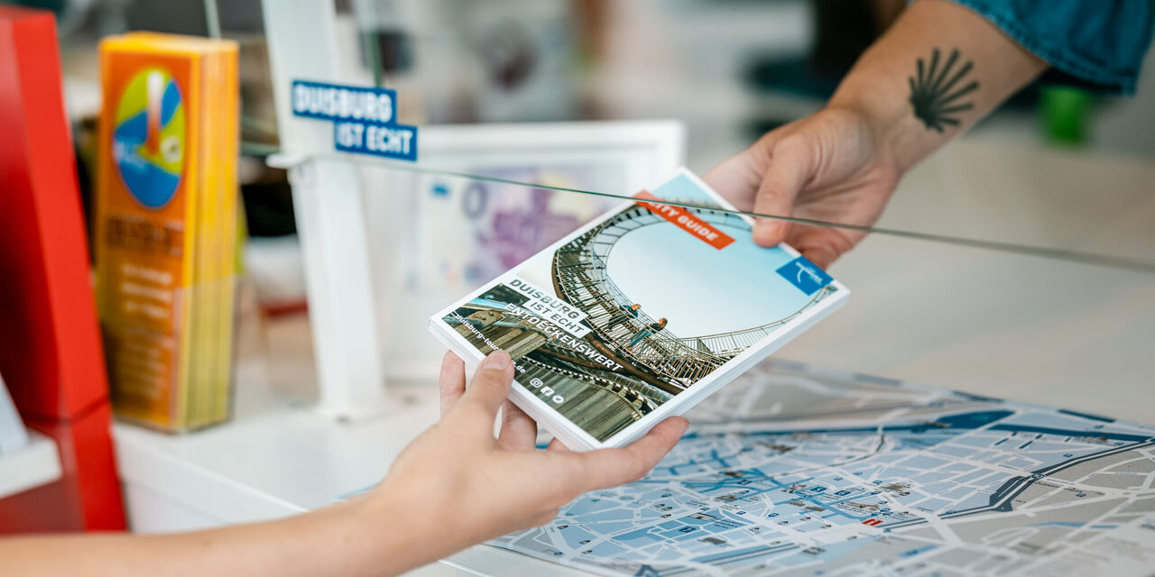 Eine Person übergibt einer weiteren Person einen City Guide, nur die Hände sind von den Personen zu sehen.