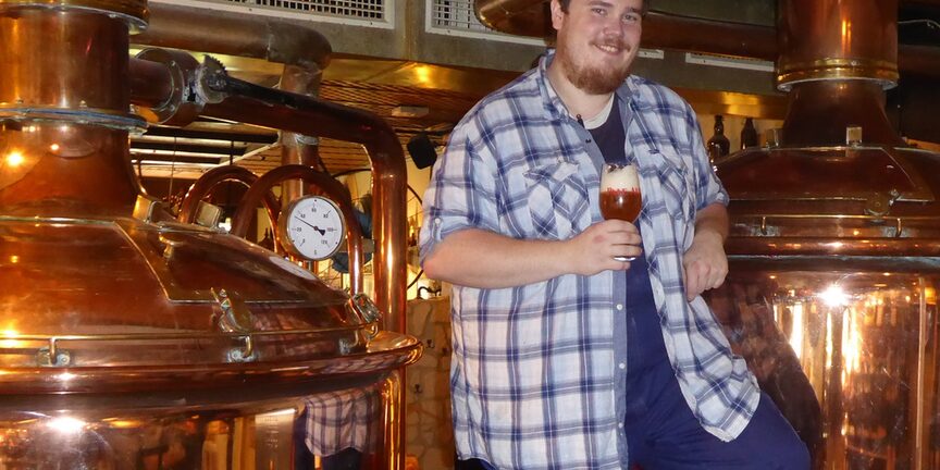 Ein Mann lehnt an einem Braukessel mit einem Glas Bier in der Hand.